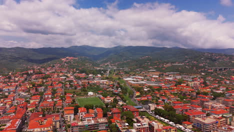 the drone moves forward over residential areas in diano marina, showing red rooftops, a soccer field, and distant mountain ranges