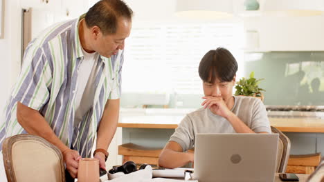 Working from home, young Asian man using laptop while older Asian man talking to him