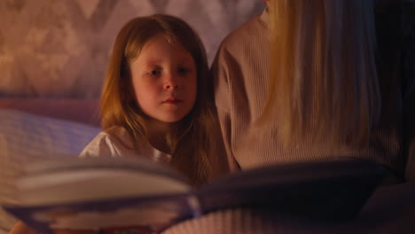 pretty child looks fairy tale book held by mother on bed