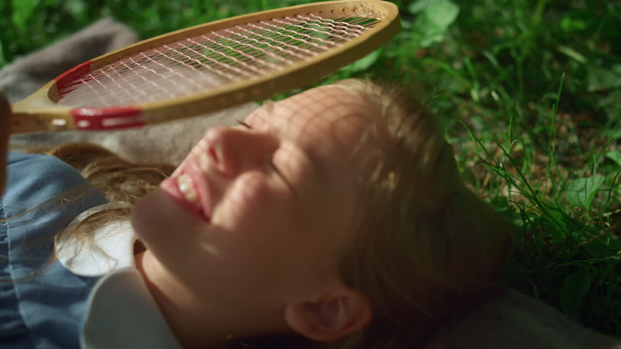 웃는 아이는 공원 클로즈업에서 담요에 누워 있습니다. 소녀는 태양에서 눈을 감고 라켓을 잡습니다.