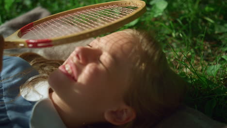 Smiling-kid-lie-blanket-in-park-closeup.-Girl-close-eyes-from-sun-hold-racket