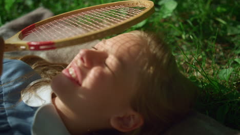 smiling kid lie blanket in park closeup. girl close eyes from sun hold racket