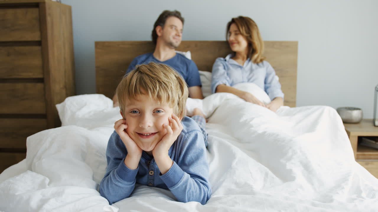 귀여운 작은 소년이 누워서 카메라를 보고 웃고 있고, 그의 부모는 그의 뒤에 있는 침대에 있는 담요 아래에 누워 있다