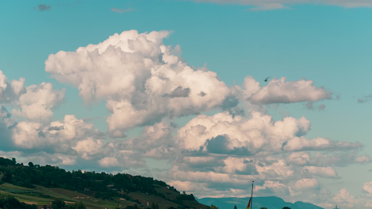 This still timelapse shows billowing cumulus clouds over lush hills at Lake Murten, Fribourg, Switzerland. The serene sky contrasts the calm lake surface.