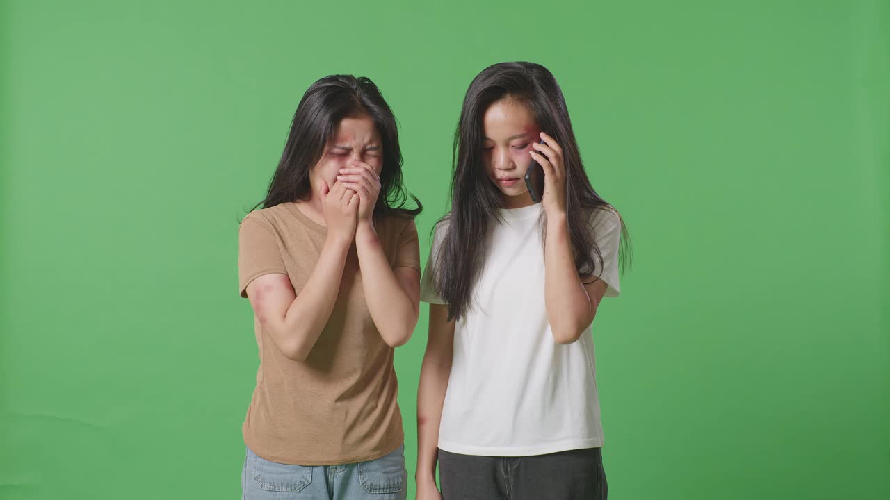 몸에 이 있는 폭력의 희생자인 젊은 아시아 여성이 눈물을 흘리는 동안 다른 여성이 스마트폰으로 이야기하면서 녹색 화면 배경 스튜디오에서 도움을 요청합니다.