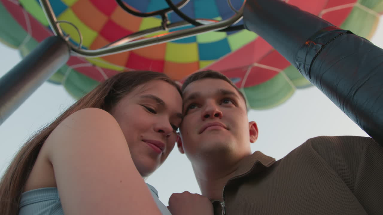 close up of young couple standing closely beneath vibrant hot air balloon canopy looking upward with thoughtful expressions as morning light reflects on their faces