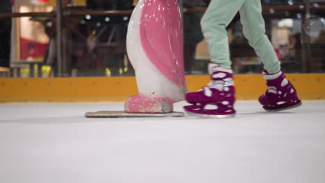 close-up of someone skating on an ice rink wearing purple skates and ash-colored trousers, accompanied by a pink and white object, with blur view of people seated outside the rink