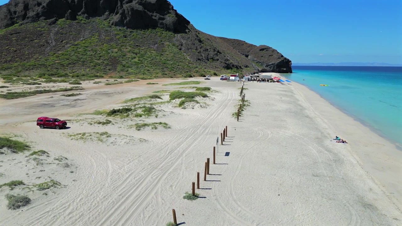 Un drone vola basso sopra la spiaggia El Tecolote vicino a Punta Shiro, seguendo una fila di pali di legno lungo la riva verso un gruppo di tende alla fine della spiaggia