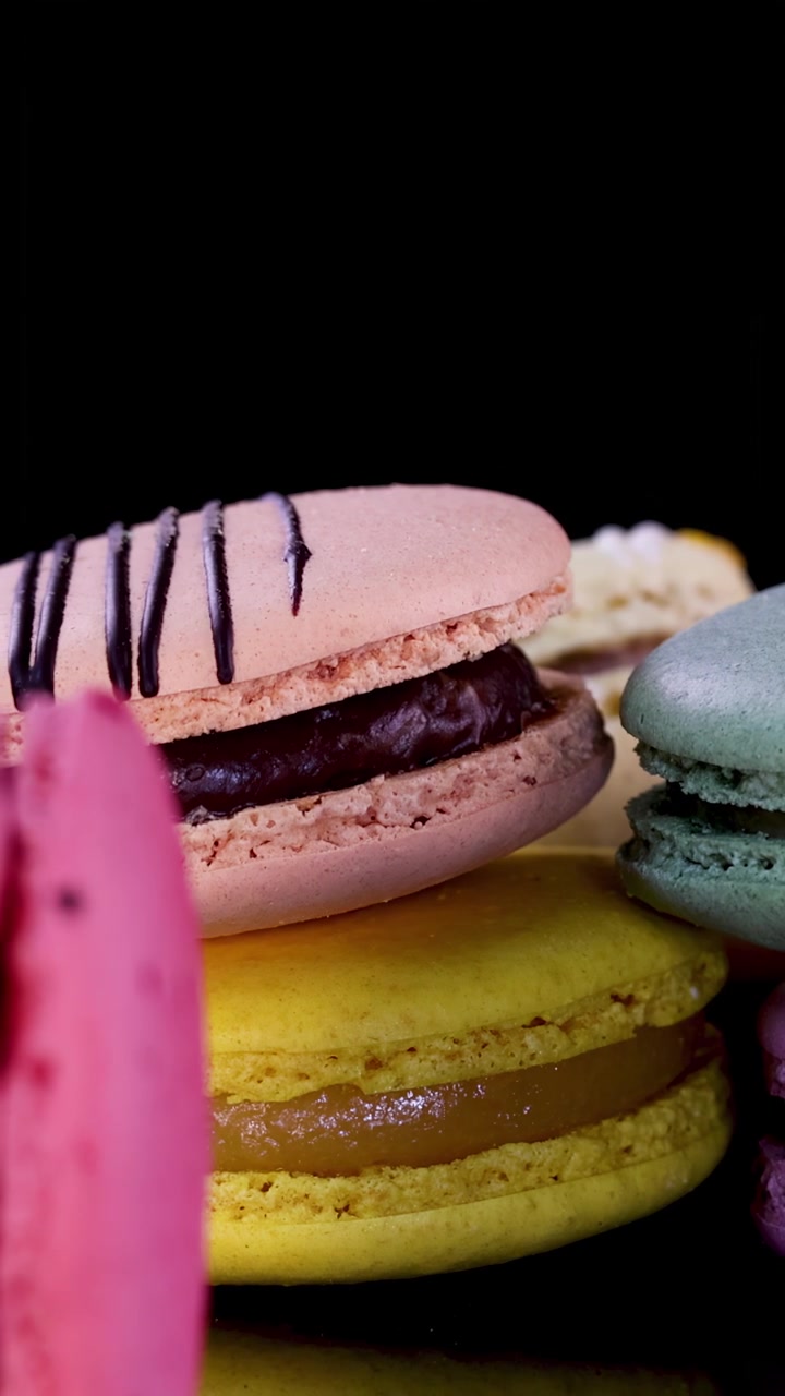 A hand carefully places assorted pastel macarons in a neat row on a glossy black surface under bright, focused lighting, highlighting vibrant dessert textures