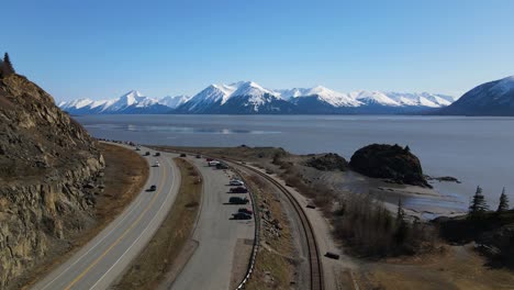 4k drone ascending above a parking lot and railroad tracks by the shoreline showcasing white capped mountains on a sunny day with a view of the bay in anchorage alaska