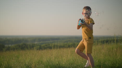 young boy wearing a yellow shirt and glasses, smiling as he holds a plastic bubble gun that releases bubbles while running through a grassy field