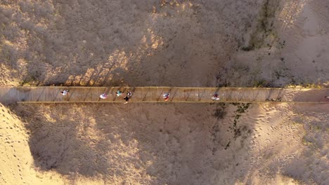 Aerial-top-down-of-group-walking-over-wooden-bridge-in-sandy-terrain-of-beach-during-sunset