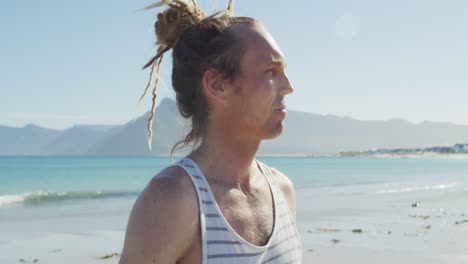 video portrait of smiling caucasian man with dreadlocks holding yoga mat on sunny beach