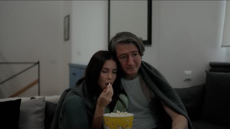 pareja sentada en el sofá tomando palomitas de maíz y viendo thriller o drama en la televisión juntos