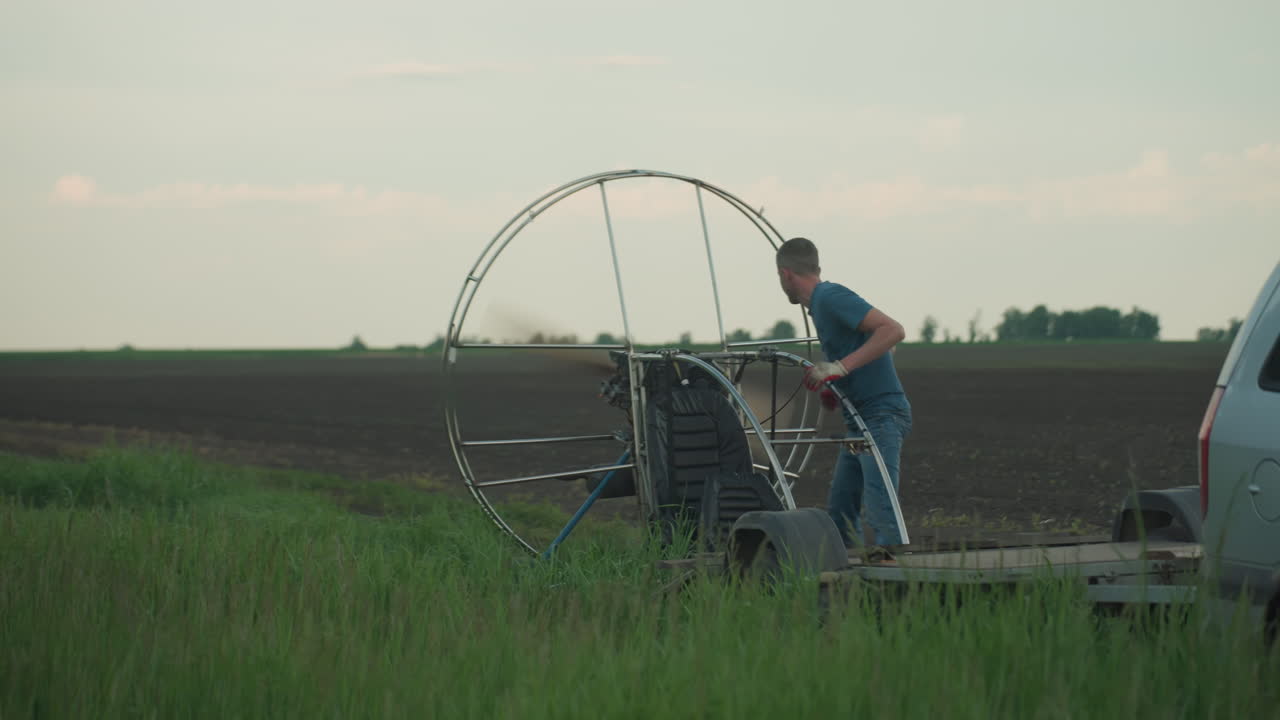 young man in blue shirt bends over lightweight paraglider on grassy field at dusk igniting engine as propeller begins spinning within circular frame against open farmland