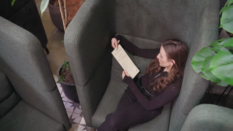 overhead view of woman with curly brown hair sitting in green cushioned booth and holding menu, absorbed in reading. indoor setting features potted plant, brick wall, and patterned tile flooring
