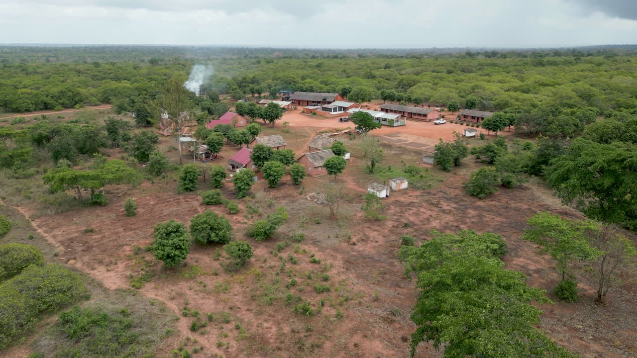Isolated rural African communal compound with simple buildings and rising smoke, surrounded by dense green African vegetation and dark clouds