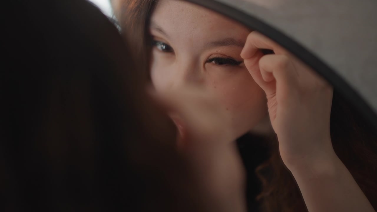 Giovane donna applica l'eyeliner mentre si guarda in uno specchio rotondo, stabilizzando l'occhio con le dita e guidando lo strumento cosmetico lungo la linea delle ciglia superiori sotto una morbida luce ad anello in un ambiente interno caldo.