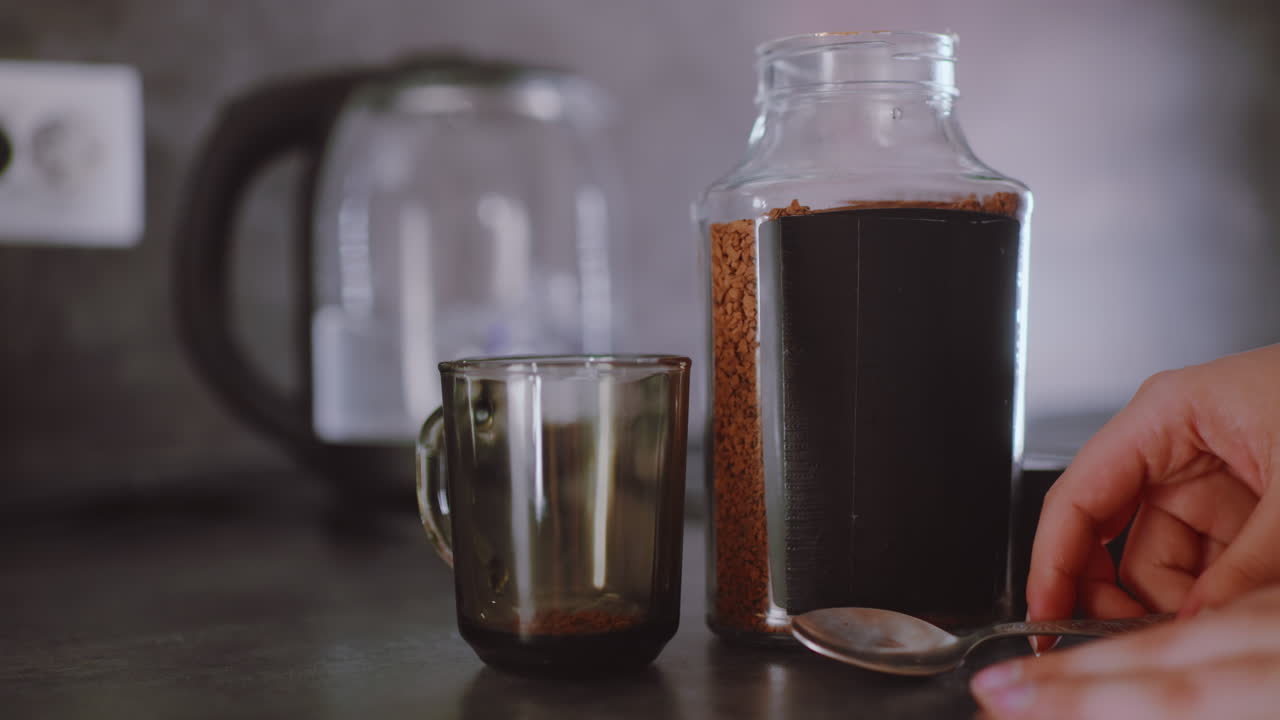 Una mano prende un cucchiaio di caffè solubile da un grande barattolo trasparente e lo versa in una tazza di vetro vicino a un bollitore elettrico, poi lascia cadere il cucchiaio sul bancone e prende il coperchio nero di un contenitore in una moderna cucina