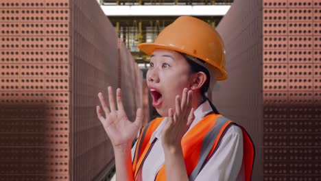 close up side view of asian female engineer with safety helmet smiling and saying wow while standing with red brick packed in stacks are stored