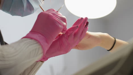 nail technician wearing pink gloves and face mask uses thin stick to gently remove excess hardener from client nail under bright lamp, showcasing attention to detail during manicure process