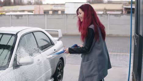 Young-woman-with-red-hair-applying-foam-on-her-silver-sportcar-with-special-jet-on-self-service-carwash