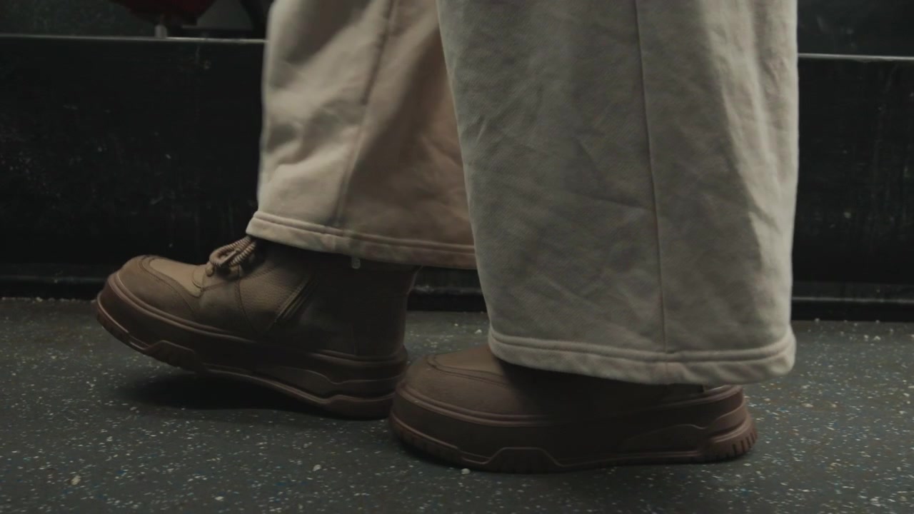 Mid-calf low-angle view showing person walking across textured floor with one heel lifted, other foot flat, and clear weight shift; light trousers drape over scuffed leather shoes in soft indoor light