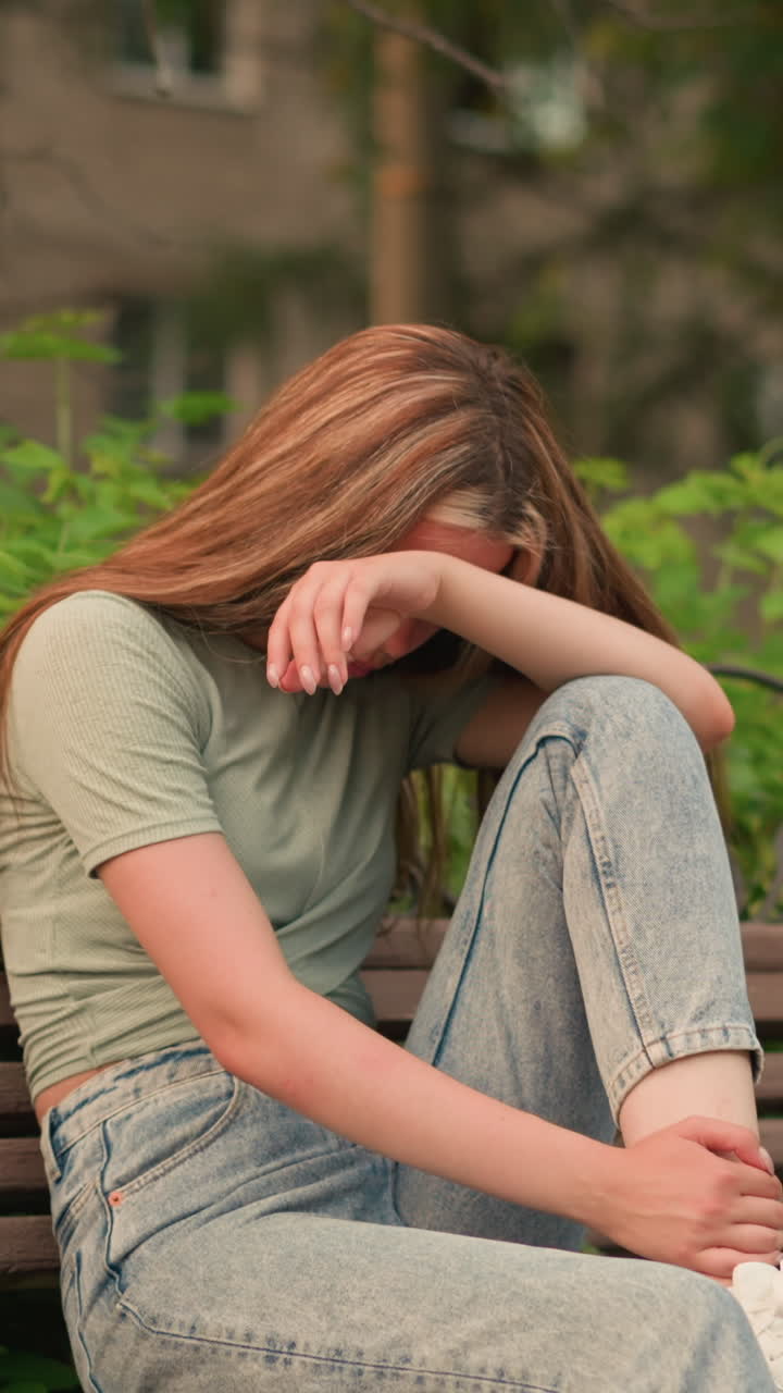 Young woman in jeans and white sneakers sits on wooden bench in park, resting her head on her arm in a reflective, somber posture, surrounded by lush greenery and trees, she appears deep in thought