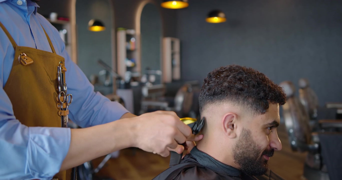 Barbeiro e cliente afro-americanos na barbearia, o barbeiro colocando a mão na cabeça, aparando um degradê com máquina.