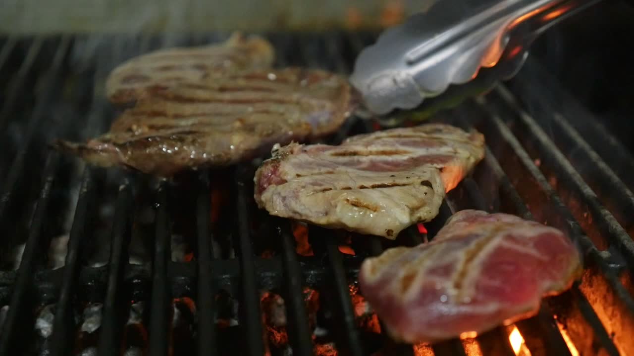 Chef using tongs to flip juicy pieces of meat on a blazing grill with flames and heavy smoke. Shows grill marks