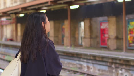 rear view of young woman waiting on railway station platform for train looking at mobile phone 1