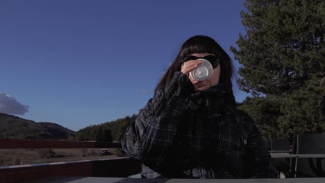 Steady-shot-of-a-young-beautiful-girl-with-brown-hair,-wearing-winter-clothes,-a-scarf-and-sunglasses,-smiling-towards-the-camera-as-she-is-picking-up-a-white-glass-to-drink-from