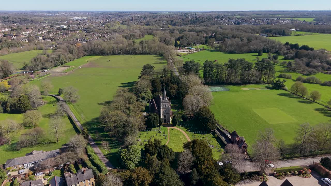 St Mary's Church drone flyover in Hertingfordbury, approaching the tower over village homes, churchyard graves, and playing field, with Hertford town and farmland beyond