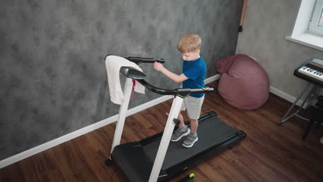 aerial view young kid walking on treadmill holding handles wiping hand on console towel draped over display casual home fitness exercise session in room with grey textured wall wooden floor seat