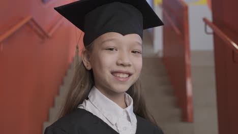 zoom in of a happy asian preschool female student in cap and gown holding graduation diploma and looking at the camera