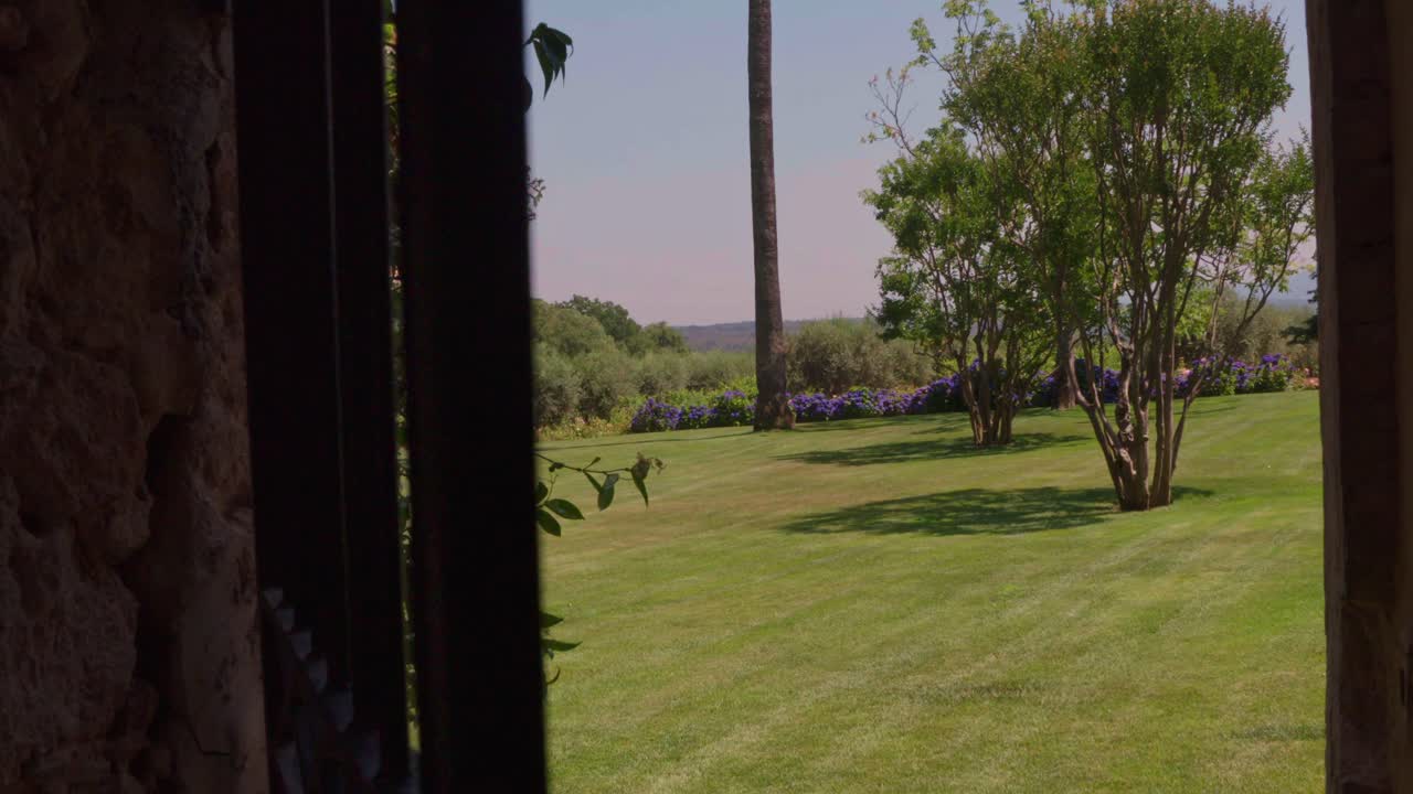 A wide shot shows an iron gate door opening to reveal a lush garden with green grass, trees, and purple flowers under a clear sky.