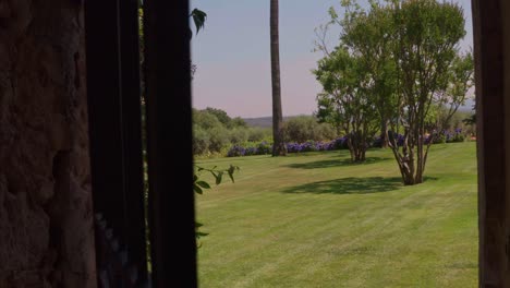 a wide shot shows an iron gate door opening to reveal a lush garden with green grass, trees, and purple flowers under a clear sky.