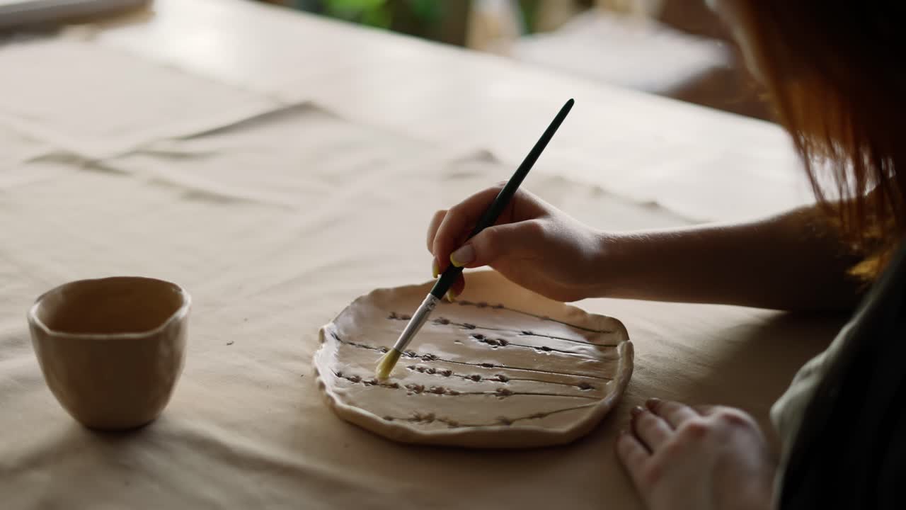 una alfarera de cabello rojo barniza un pedazo de arcilla con un pincel especial