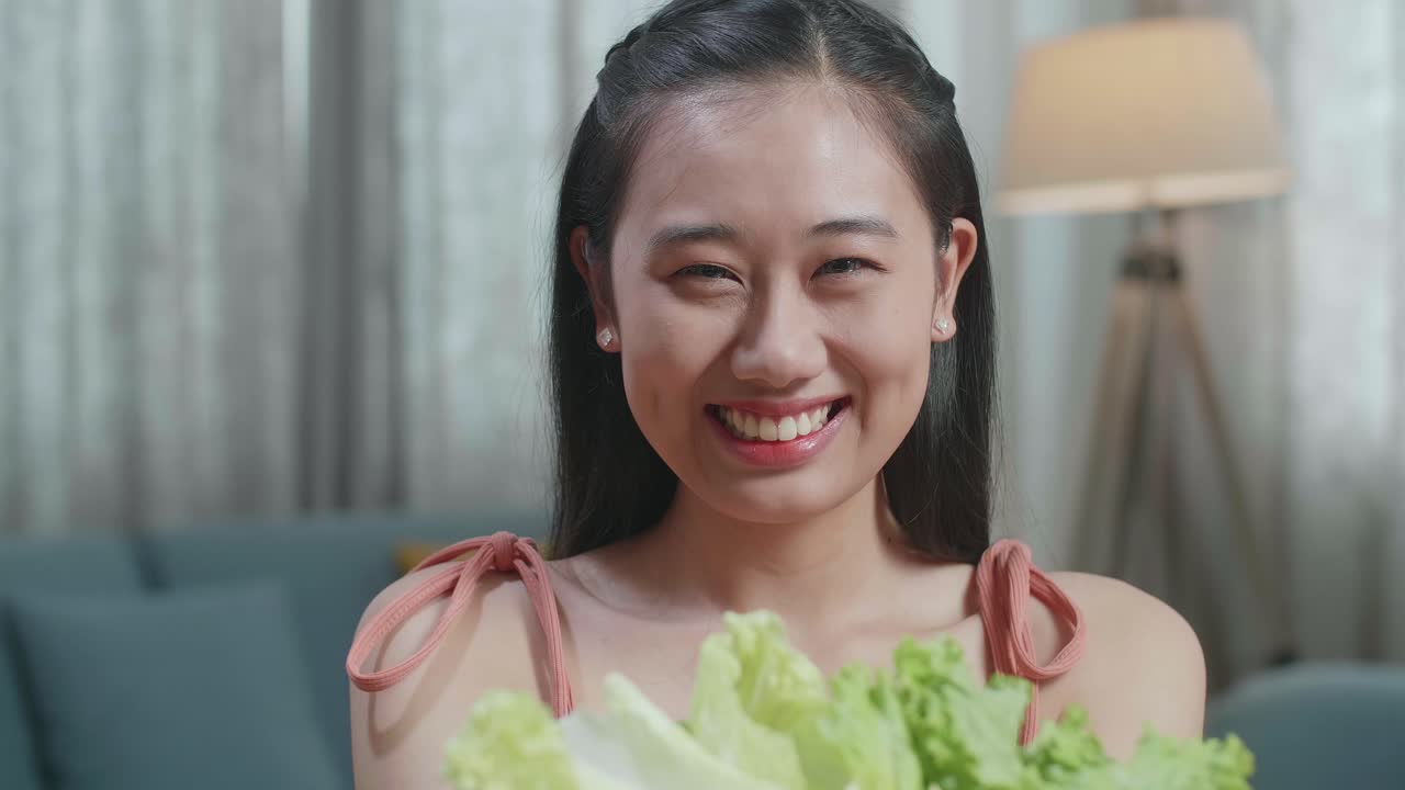 미소 짓는 아시아 여성이 집에 앉아있는 동안 건강한 음식의 접시를 카메라에 보여주고 있습니다.