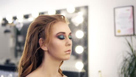 impresionante modelo caucásico con cabello largo retorcido y ojos de camaleón maquillaje. estilista acabando el maquillaje con spray. espejo de salón en el fondo