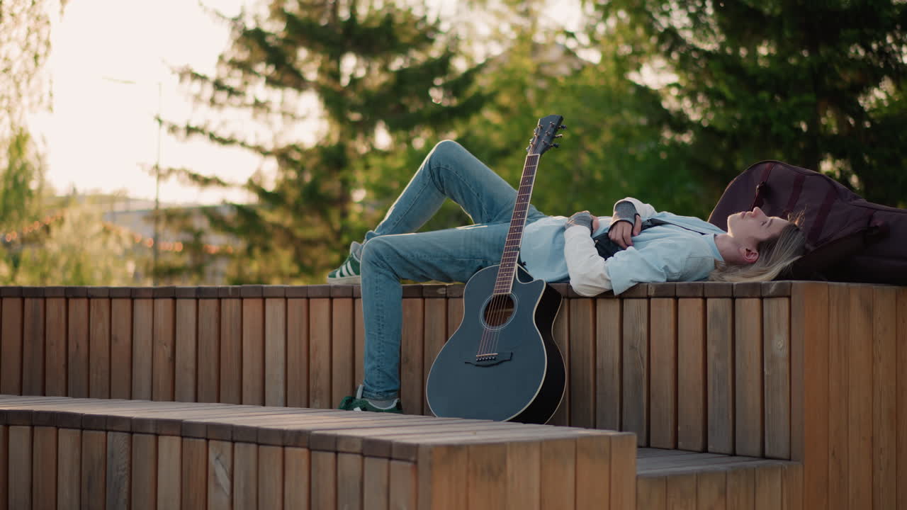 Akustikgitarrist im Sonnenuntergang ruht sich auf einer Bank aus, Rucksack in der Nähe, Gitarre lehnt an der Holzbank, warmes Gegenlicht durch Kiefern, lässige Jeans und Hoodie, entspannter Songwriter macht Pause, verträumt