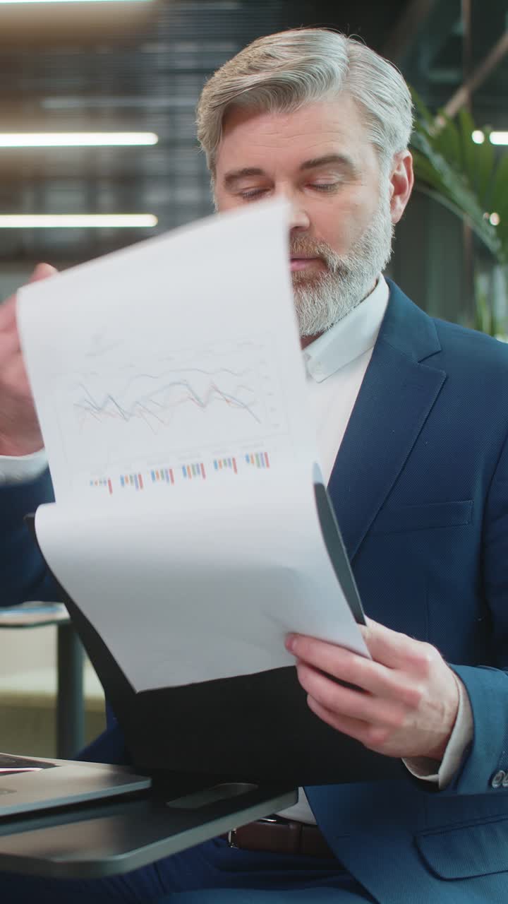 Happy male entrepreneur in suit analyzing graph documents reports and using laptop in office lobby
