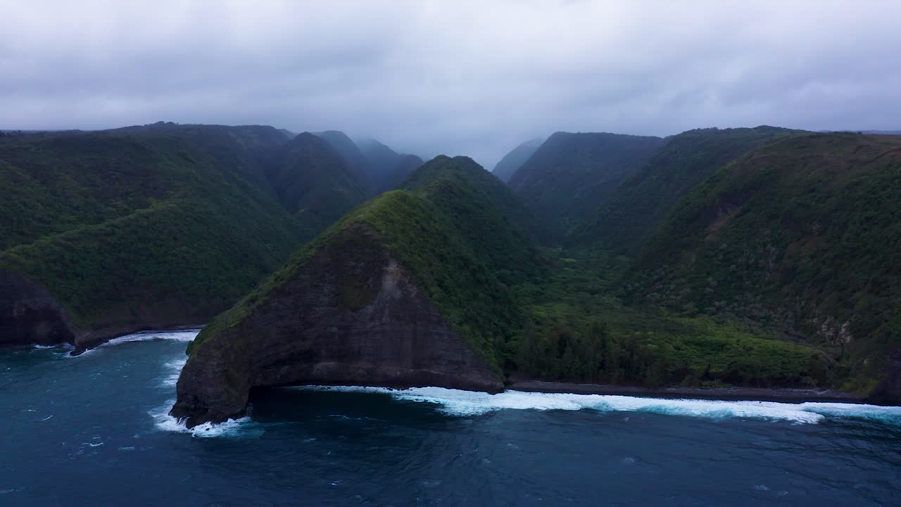 A dramatic rocky headland juts into the turquoise Pacific Ocean at Polulu Valley, Hawaii, framed by steep green cliffs and misty mountain ridges in the distance.