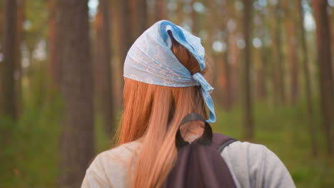 back view of young woman hiking through lush forest, wearing a blue bandana and backpack, surrounded by vibrant greenery and tall trees with warm glow the background