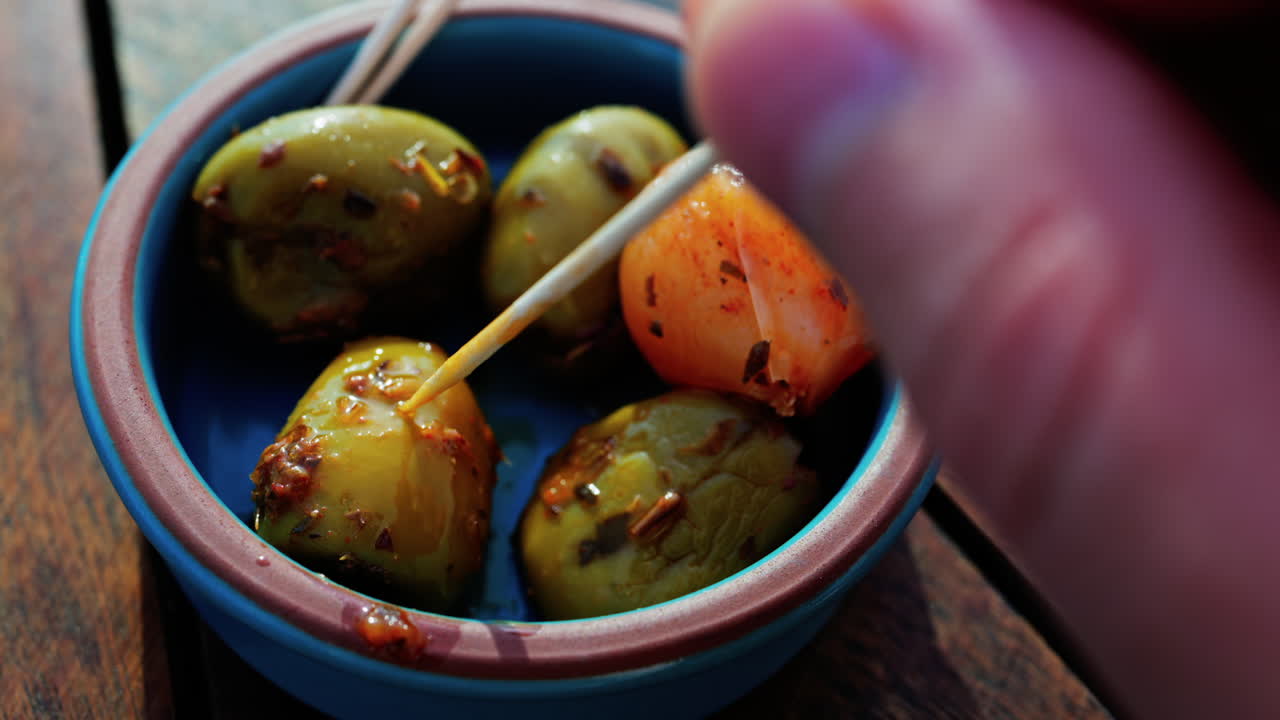Close up of a hand picking up a green olive with a toothpick on a small bowl on a table