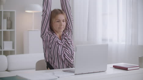 student woman is learning online with laptop and copybook at home stretching tired muscles of back and neck