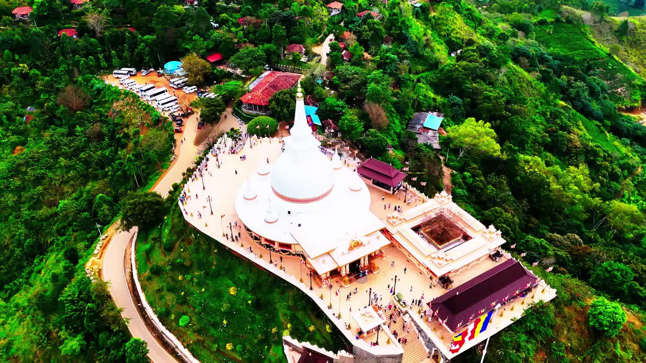 A Sri Lankan hilltop temple with a striking white stupa, surrounded by lush hills, pathways, visitors, and colorful flags, blending spirituality with natural beauty.