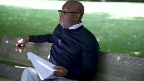 professional man taking a relaxing break on a park bench, vaping and reading papers on a sunny day