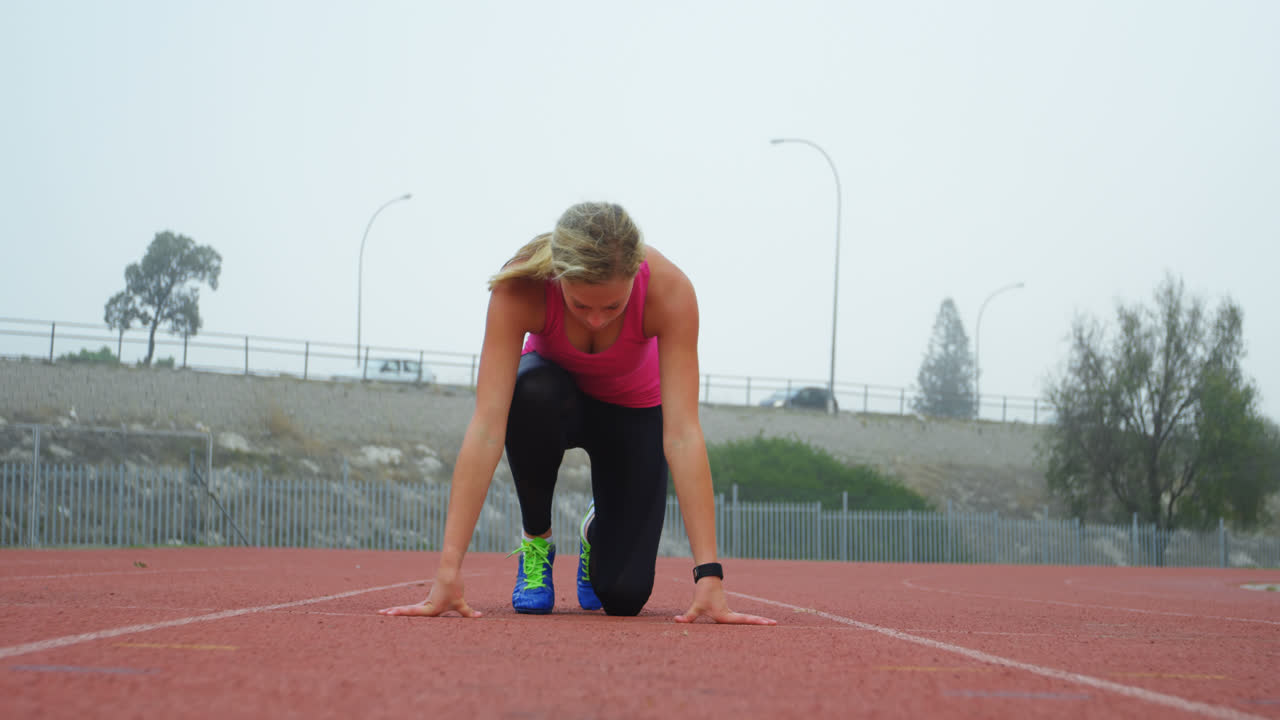 vista frontal de una atleta caucásica tomando la posición de salida en la pista de carrera en un lugar deportivo 4k