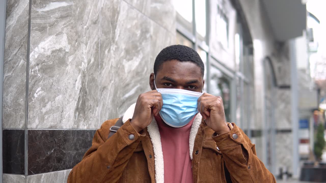 Black man with medical mask and shoulder backpack stands outdoor near the office center